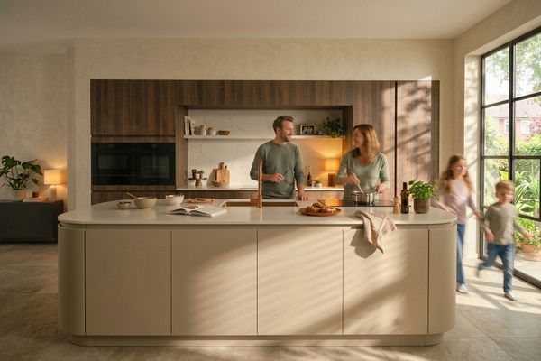 Een gezin in een beige keuken: ouders koken en de twee kinderen rennen langs een groot raam.