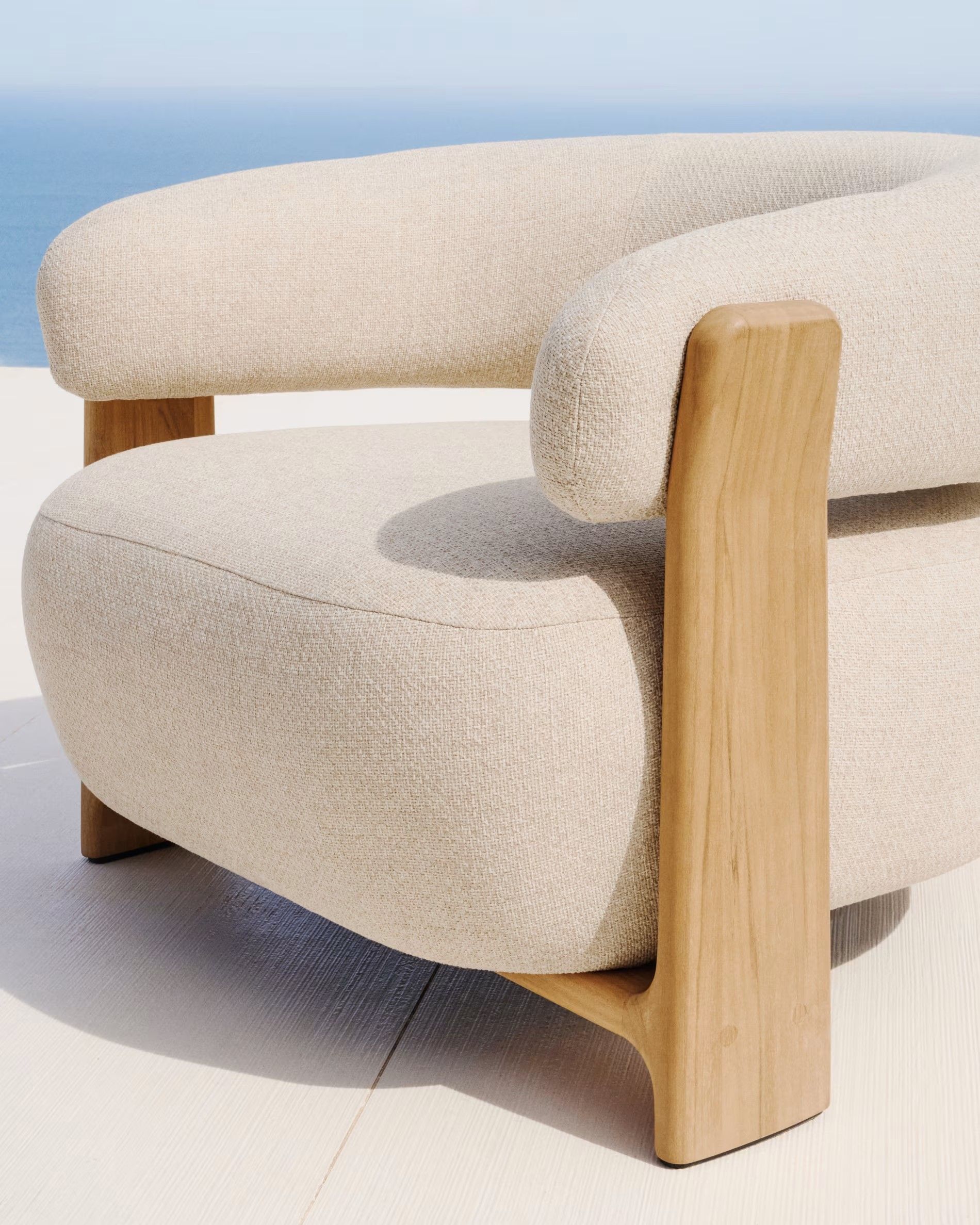 Loungefauteuil Granite in beige stof met houten armleuningen en poten, tegen een blauwe horizon.