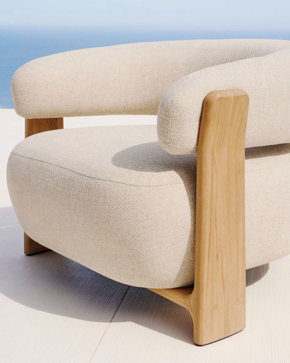 Loungefauteuil Granite in beige stof met houten armleuningen en poten, tegen een blauwe horizon.