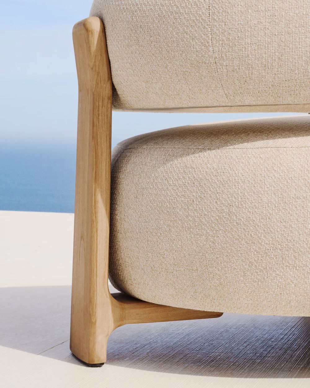 Loungefauteuil GRANITE met beige stoffen bekleding en massief teakhouten frame tegen een achtergrond van de zee.