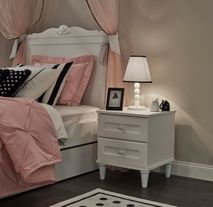 Wit houten nachtkastje LORA met twee lades naast een opgemaakt bed.