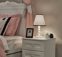 Wit houten nachtkastje LORA met twee lades naast een wit bed in een kinderkamer.