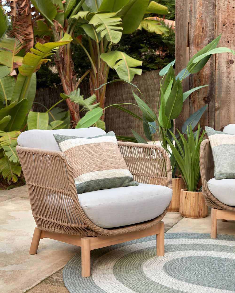 Loungefauteuil CATALINA van beige wicker met lichtgrijze kussens, een gestreept sierkussen, en acaciahouten poten.