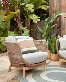 Loungefauteuil CATALINA van beige wicker met lichtgrijze kussens, een gestreept sierkussen, en acaciahouten poten.