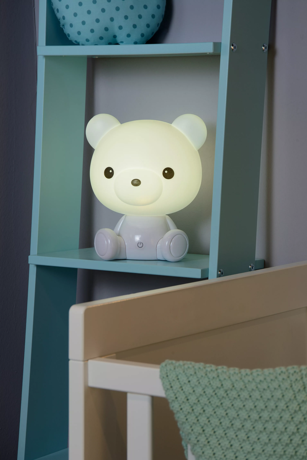 Witte kunststof teddybeer tafellamp op een blauwe plank, met een babybedje op de voorgrond.