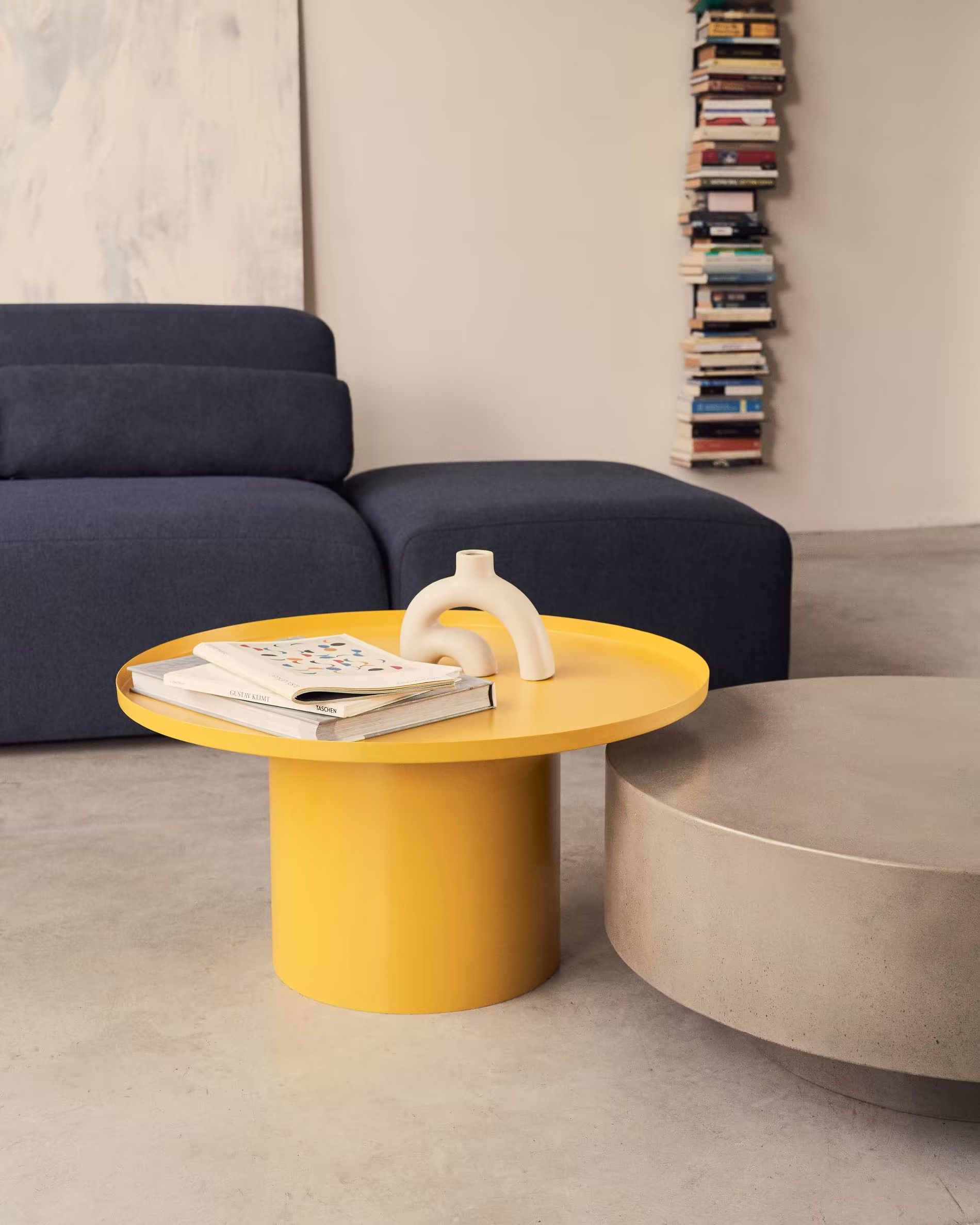 Ronde gele metalen salontafel FLEKSA met wit ornament en boeken, met donkerblauwe bank en cementkleurige tafel.