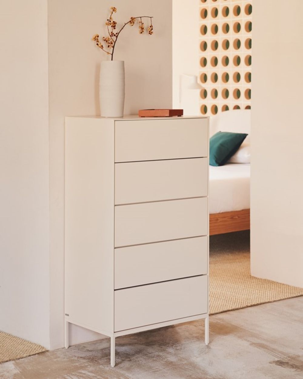 Witte houten commode met vijf lades en metalen poten, met vaas en doosje erop, in een slaapkamer.
