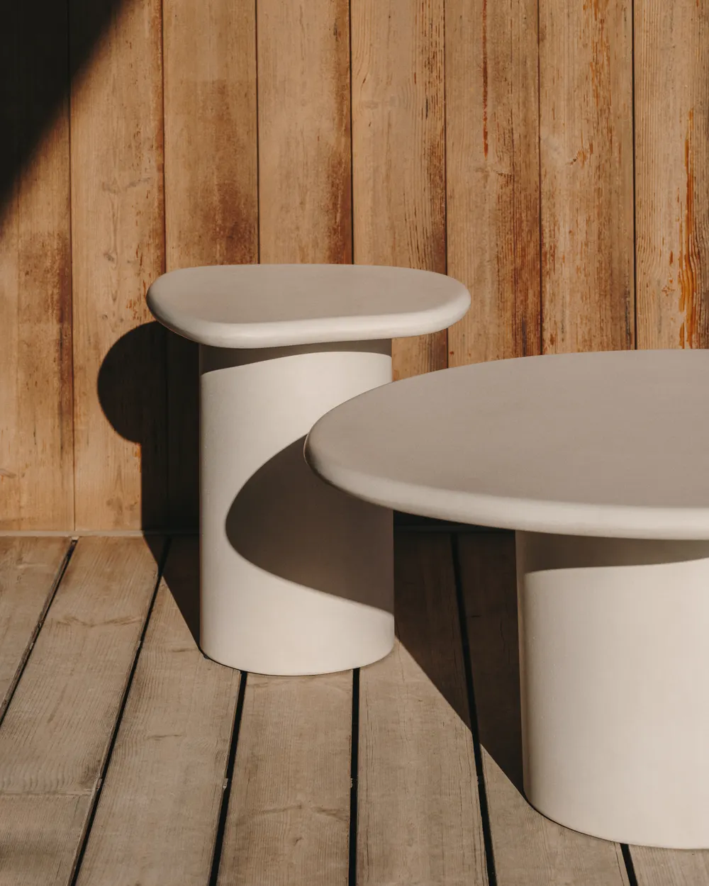 Twee beige bijzettafels van cement op een houten terras tegen een houten wand.