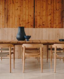 Lange houten tafel met houten stoelen en donkerblauwe vazen en schalen, tegen een houten wand.