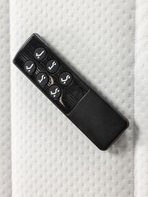 Zwarte kunststof afstandsbediening met grijze knoppen voor een elektrische boxspring, liggend op een wit matras.