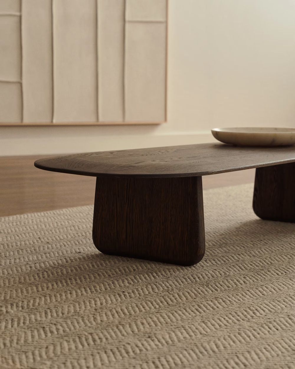 Salontafel PIRITA, ovaal blad van donker massief eikenhout op twee poten, staat op beige vloerkleed.