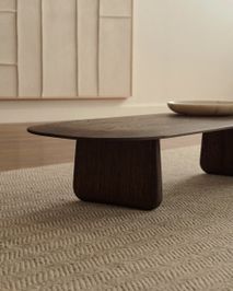 Salontafel PIRITA, ovaal blad van donker massief eikenhout op twee poten, staat op beige vloerkleed.