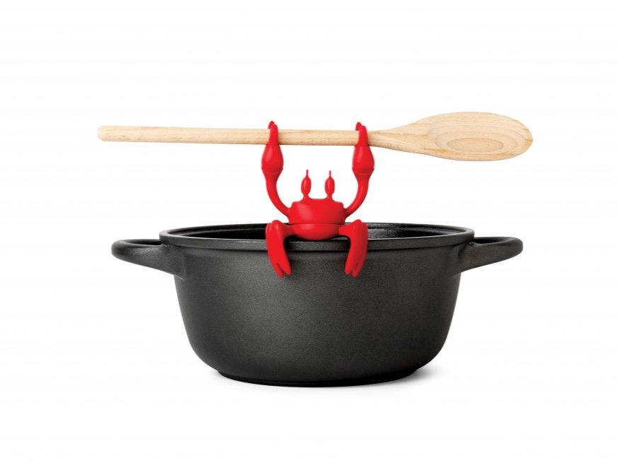 Keukenhulp RED KRAB: rode siliconen krab houdt een houten lepel op een zwarte metalen pan.