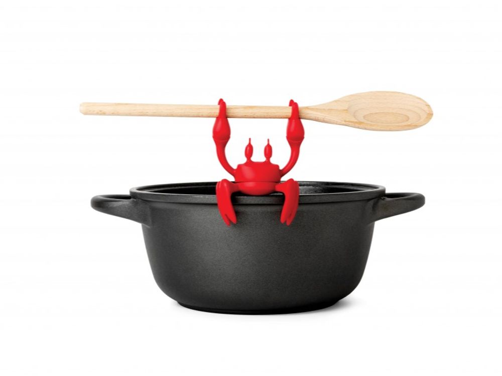 Keukenhulp RED KRAB: rode siliconen krab houdt een houten lepel op een zwarte metalen pan.