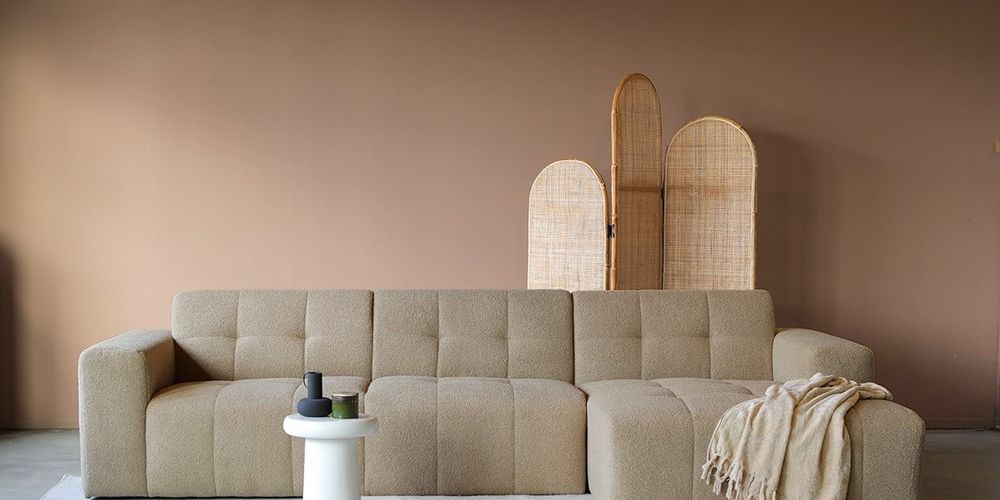 Moderne lichte hoekbank tegen een taupe muur met roomwit bijzettafeltje, decoratie en afscheidingsscherm.