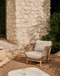 Loungefauteuil CATALINA van beige rotan en acaciahout met lichte kussens, staand op een bruin kleed.